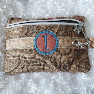 May include: A brown paisley fabric zippered pouch with a blue and pink embroidered circle containing the letter "I". The pouch has a silver zipper pull and a silver clasp.