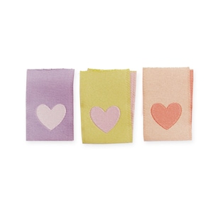 Puede incluir: Tres etiquetas de tela en colores pastel: lavanda, amarillo y melocotón. Cada etiqueta presenta un diseño en forma de corazón en un color contrastante. Estas etiquetas probablemente se utilizan para proyectos de costura o manualidades.