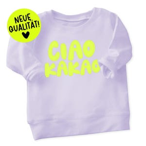 Könnte beinhalten: Ein helllila Langarm-Sweatshirt mit den Worten "CIAO KAKAO" in leuchtend gelben Buchstaben. Ein gelber Kreis mit den Worten "NEUE QUALITAT!" und einem schwarzen Herz befindet sich in der oberen linken Ecke.