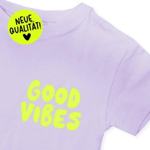 Pode incluir: Camiseta lilás clara com as palavras "GOOD VIBES" em letras amarelas brilhantes. Um adesivo amarelo neon com o texto "NEUE QUALITÄT!" e um coração preto está no canto superior esquerdo.
