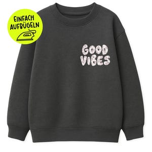 Könnte beinhalten: Dunkelgraues Sweatshirt mit den Worten "GOOD VIBES" in weißen Buchstaben auf der Brust. Ein gelber Kreis mit deutschem Text und einem Bügeleisen befindet sich oben links. Das Sweatshirt hat einen Rundhalsausschnitt und lange Ärmel.