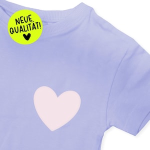 Könnte beinhalten: Ein helllila T-Shirt mit einem großen, hellrosa Herz auf der Vorderseite. Ein gelber Aufkleber mit dem Text "NEUE QUALITÄT!" und einem schwarzen Herz befindet sich oben links. Das Shirt hat kurze Ärmel.
