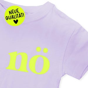 Könnte beinhalten: Helllila T-Shirt mit dem Wort "nö" in leuchtendem Gelb, mit zwei gelben Punkten über dem "ö". Ein gelber Kreis mit dem Text "NEUE QUALITÄT!" und einem schwarzen Herz befindet sich oben links.