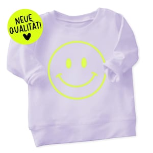 Könnte beinhalten: Ein helllila Sweatshirt mit einem großen neongelben Smiley-Gesicht. Ein neongelber Kreis mit dem Text "NEUE QUALITAT!" befindet sich in der oberen linken Ecke. Das Sweatshirt hat lange Ärmel und einen Rundhalsausschnitt.