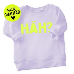 Könnte beinhalten: Ein helllila Sweatshirt mit dem Wort "HÄH?" in leuchtend gelben Buchstaben. Ein gelber Kreis mit den Worten "NEUE QUALITÄT!" und einem schwarzen Herz befindet sich in der oberen linken Ecke. Das Sweatshirt hat lange Ärmel.