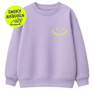 Könnte beinhalten: Ein helllila Sweatshirt mit Rundhalsausschnitt und gerippten Bündchen und Saum. Das Sweatshirt hat ein gelbes Smiley-Gesicht-Design mit den Worten "always smile" und einen gelben Kreis mit den Worten "einfach aufbügeln" und einem Bügeleisensymbol.