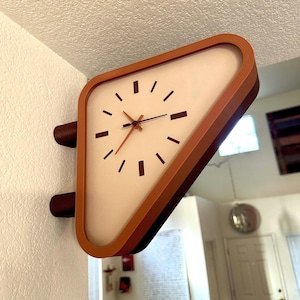 May include: A triangular wall-mounted clock with a burnt orange frame and a white face. The clock features dark brown hour markers and thin, matching brown hands. It is attached to the wall by two cylindrical dark brown mounting brackets.