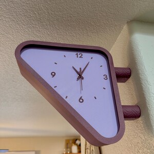 May include: A triangular wall-mounted clock with a dark purple frame and a light lavender face. The clock features dark purple hands and numerals at the 3, 6, 9, and 12 o'clock positions. It is attached to the wall by two textured purple mounts.