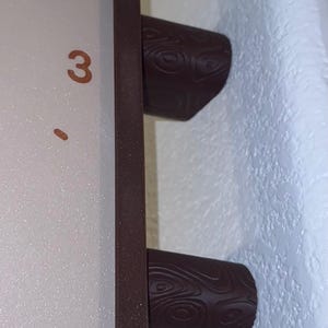 May include: A rectangular clock with a light-colored face featuring the number 3 in a brown font. The clock has a dark brown frame and two cylindrical feet with a textured, swirling wood-grain pattern on the exterior surface.