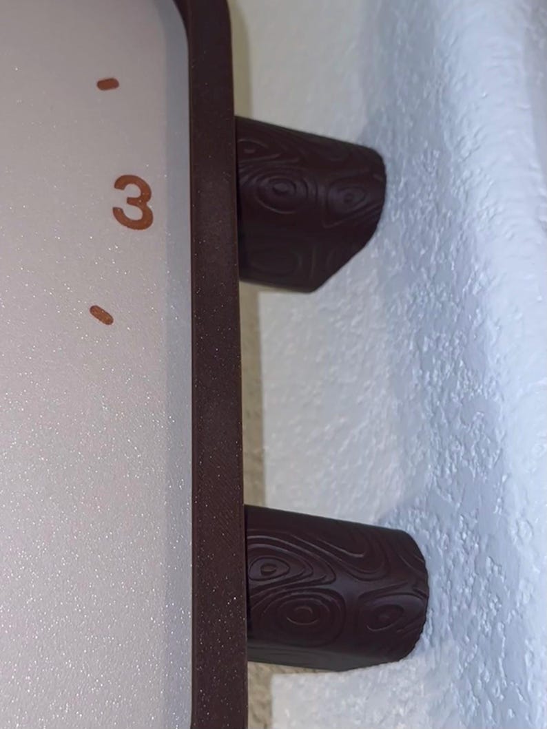May include: A rectangular clock with a light-colored face featuring the number 3 in a brown font. The clock has a dark brown frame and two cylindrical feet with a textured, swirling wood-grain pattern on the exterior surface.