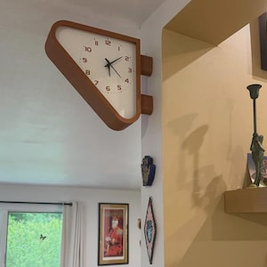 May include: A double-sided wall clock with a triangular wooden frame and a white face featuring black numerals and hands. The clock is mounted on a corner wall, extending outward with a distinct geometric shape and a clean, minimalist aesthetic.