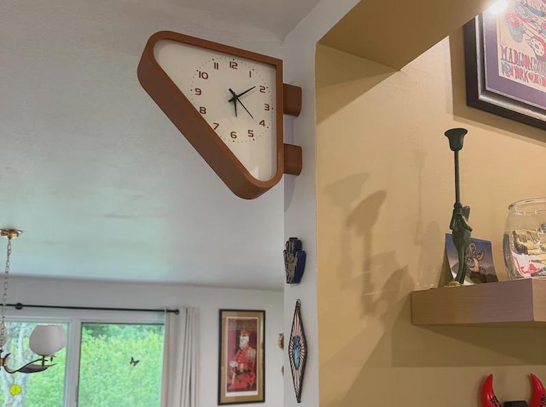 May include: A double-sided wall clock with a triangular wooden frame and a white face featuring black numerals and hands. The clock is mounted on a corner wall, extending outward with a distinct geometric shape and a clean, minimalist aesthetic.
