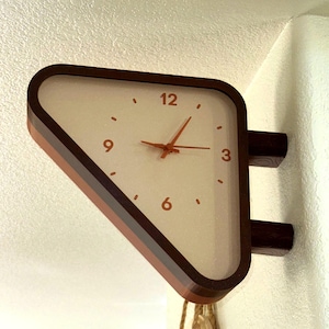 May include: A triangular wall-mounted clock with a dark brown frame and a cream-colored face. The clock features copper-colored hands and numerals for 3, 6, 9, and 12, with small dots marking the remaining hours. It is attached to the wall by two brackets.