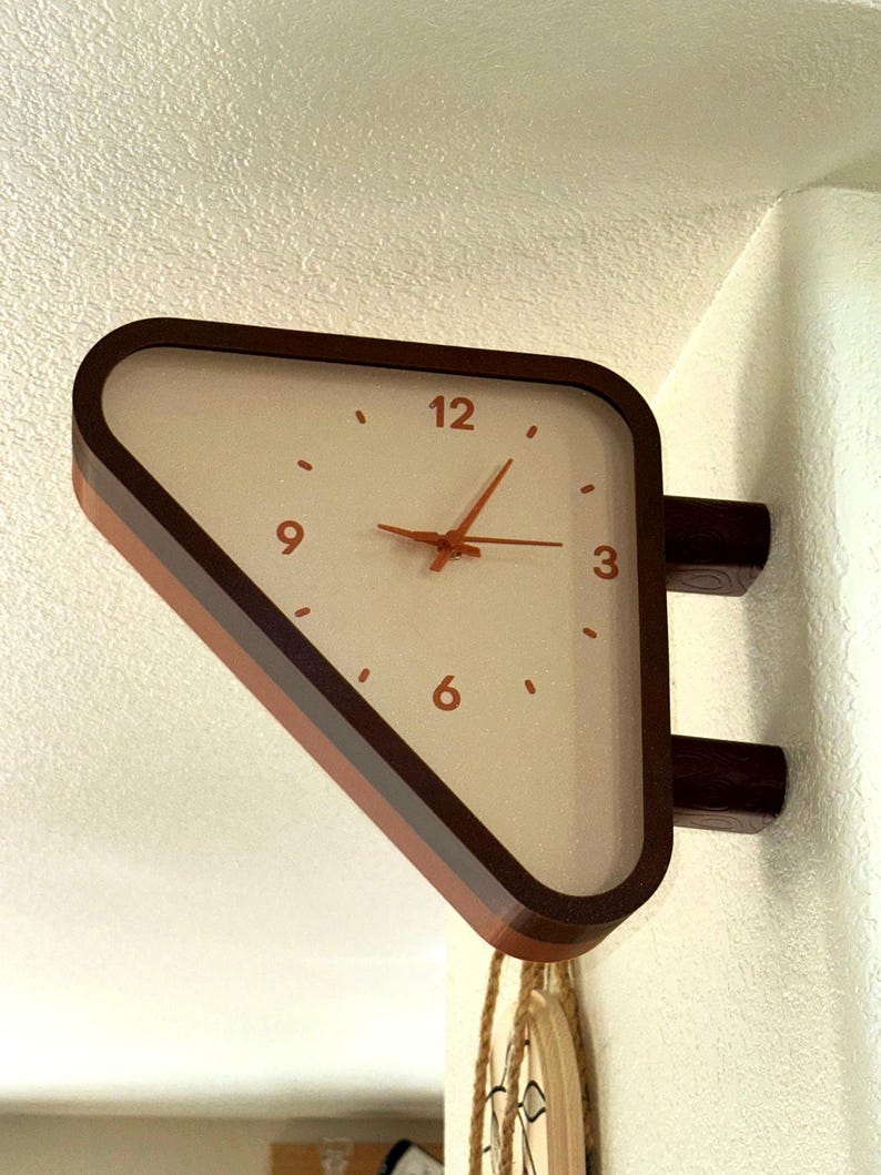 May include: A triangular wall-mounted clock with a dark brown frame and a cream-colored face. The clock features copper-colored hands and numerals for 3, 6, 9, and 12, with small dots marking the remaining hours. It is attached to the wall by two brackets.