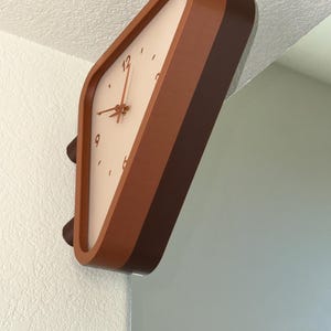 May include: A wall-mounted clock with a rounded triangular frame in shades of brown. The clock face is off-white with brown numerals at the 12, 3, 6, and 9 positions, featuring thin brown hour, minute, and second hands centered on the dial.