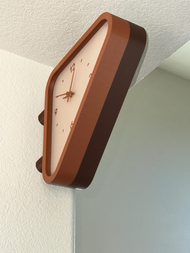 May include: A wall-mounted clock with a rounded triangular frame in shades of brown. The clock face is off-white with brown numerals at the 12, 3, 6, and 9 positions, featuring thin brown hour, minute, and second hands centered on the dial.