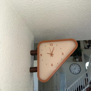 May include: A triangular wall-mounted clock with a terracotta-colored frame and a cream-colored face. The clock features dark hands and the numbers 3, 6, 9, and 12 printed in a dark color. It is attached to the wall by two cylindrical mounts.