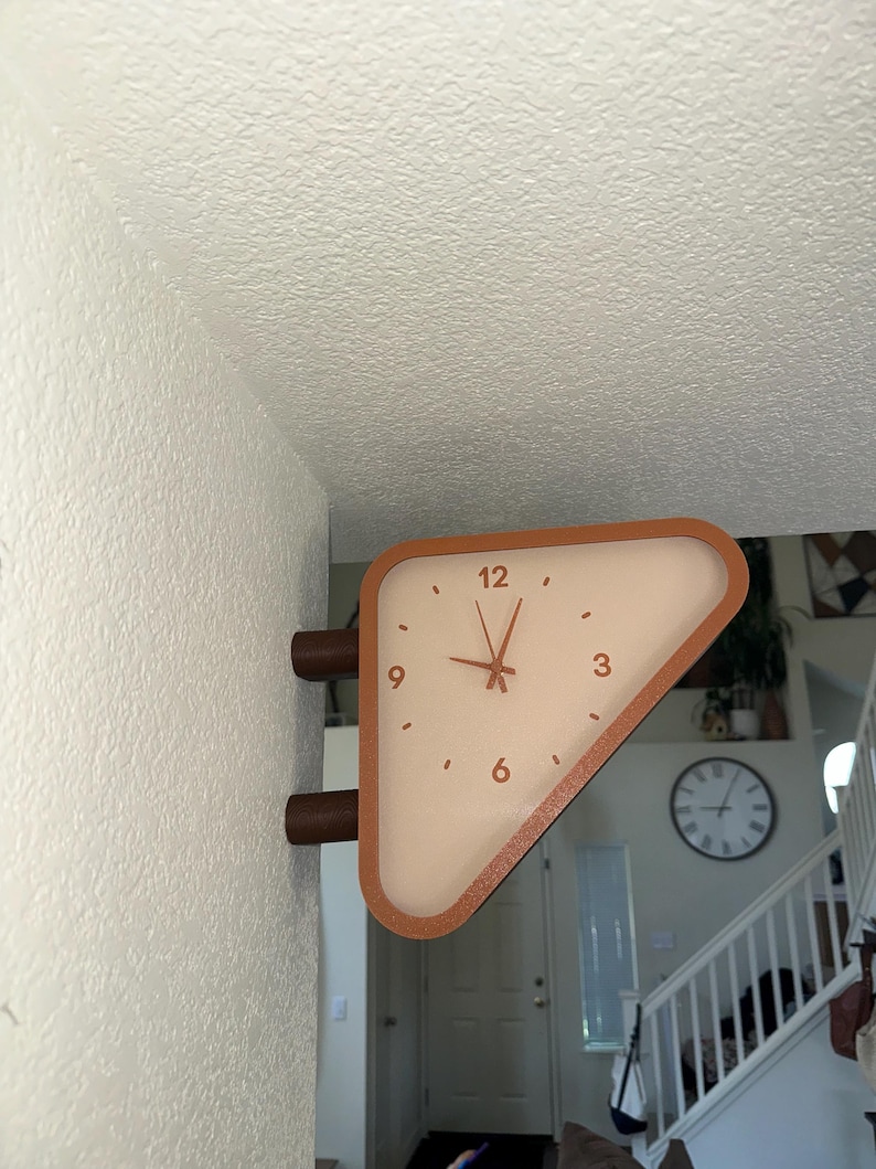 May include: A triangular wall-mounted clock with a terracotta-colored frame and a cream-colored face. The clock features dark hands and the numbers 3, 6, 9, and 12 printed in a dark color. It is attached to the wall by two cylindrical mounts.
