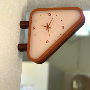 May include: A triangular wall-mounted clock with a burnt orange frame and a translucent white face. The clock features dark orange hour, minute, and second hands, with the numbers 3, 6, 9, and 12 printed in a dark orange font on the face.