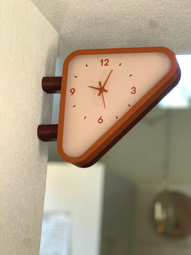 May include: A triangular wall-mounted clock with a burnt orange frame and a translucent white face. The clock features dark orange hour, minute, and second hands, with the numbers 3, 6, 9, and 12 printed in a dark orange font on the face.