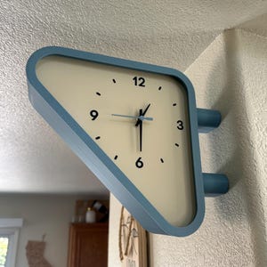 May include: A triangular wall clock with a light blue frame and a cream-colored face. The clock features black numerals at the 3, 6, 9, and 12 o'clock positions, with black hour and minute hands and a light blue second hand, mounted to the wall.