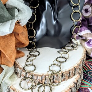 May include: A long necklace composed of numerous interlocking circular metal rings in an antique brass finish. The chain features a repeating pattern of varying ring sizes, creating a continuous, fluid design that hangs in a layered loop.