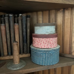May include: A stack of three round, lidded boxes with patterned paper exteriors. The top box is light blue and white gingham, the middle box is red with a white floral print, and the bottom box is dark blue with a small white geometric pattern.