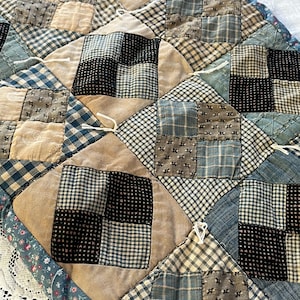 May include: Close-up of a handmade quilt with a patchwork design. The quilt features squares of various fabrics in beige, black, and blue, with a floral border. The quilt is a mix of patterns and textures.