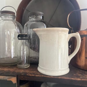 May include: A collection of vintage kitchenware, including a small white ceramic pitcher with a handle, several glass jars with metal lids, and a copper pot. The items are arranged on a wooden shelf, creating a rustic aesthetic.