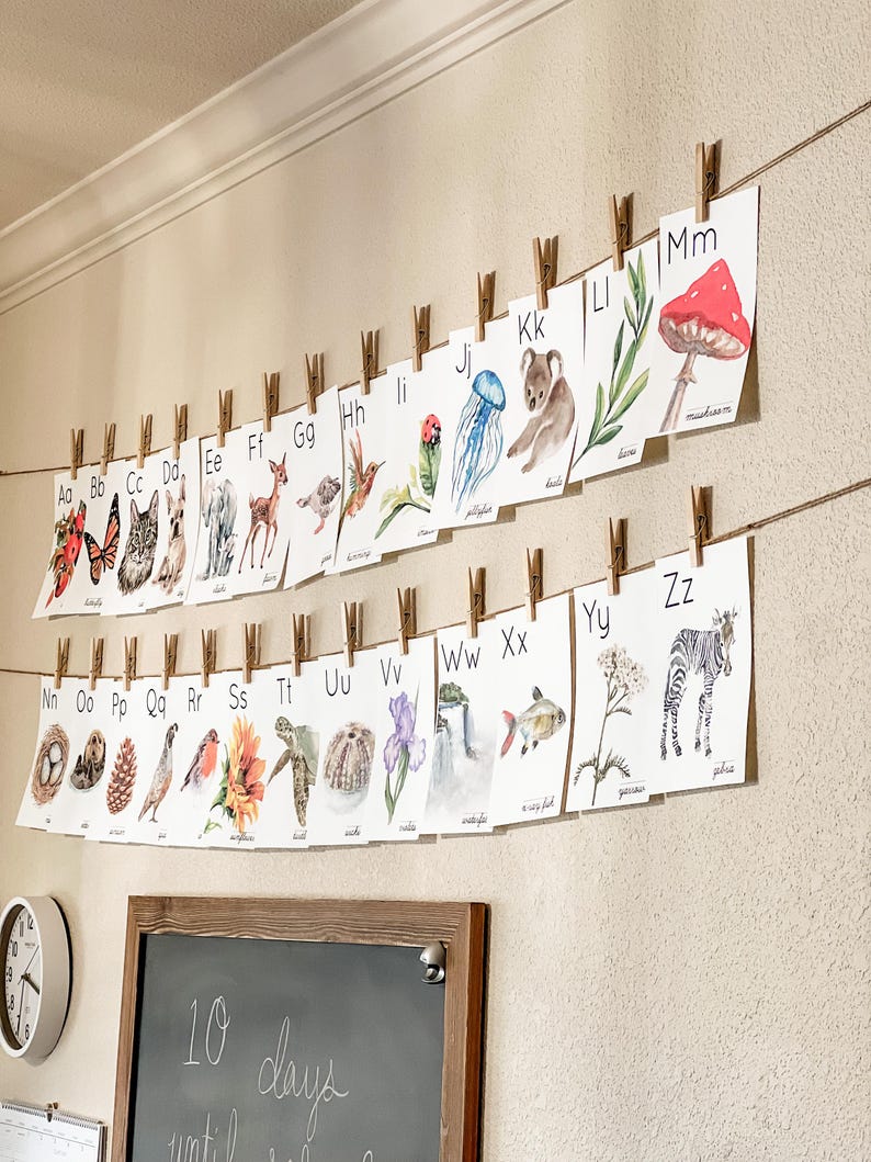 May include: A watercolor alphabet set, each letter displayed on a card with a corresponding illustration. The cards are suspended by clothespins on twine against a neutral wall. The images include a butterfly, cat, deer, and zebra.