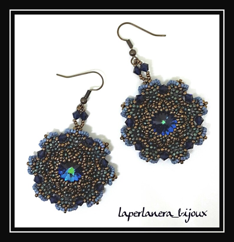 May include: A pair of blue and bronze beaded earrings with a dark blue crystal in the center of each earring. The earrings are hanging from silver hooks. The text "laperlanera_bijoux" is written in black ink at the bottom of the image.