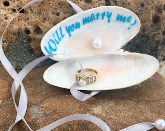 May include: A seashell with the words "Will you marry me?" written in blue on the inside. A gold ring with a diamond is inside the shell, along with a pearl. A grey ribbon with two pearls is on a brown rock.