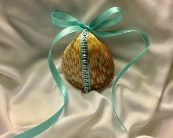 May include: A seashell with a light brown and white pattern is decorated with a turquoise ribbon and a line of rhinestones. The seashell is on a white fabric background.