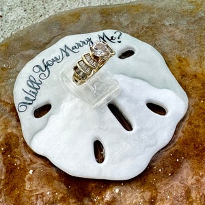 Puede incluir: Un anillo de compromiso de oro con una gran piedra central redonda y diamantes baguette en la banda, presentado sobre un dólar de arena blanco. El dólar de arena tiene la inscripción "Will You Marry Me?"