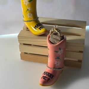 Puede incluir: Dos sujetadores de anillos con forma de bota de vaquero de cerámica. Uno es amarillo con flores blancas y el texto "MARRY ME". El otro es rosa con flores blancas y el texto "MARRY ME?". Ambos tienen una banda de diamantes de imitación.