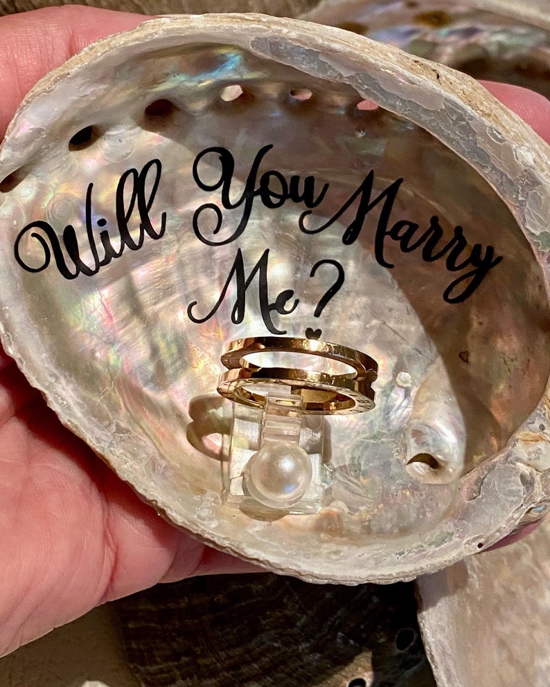 May include: A seashell ring holder with the words "Will You Marry Me?" written inside. A gold ring is displayed on the holder with a pearl in the center.