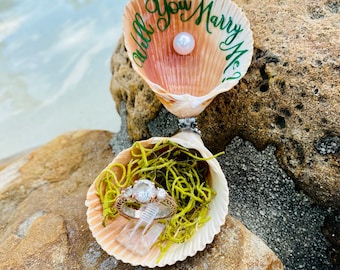 May include: A seashell ring box with the words "Will You Marry Me?" written in green calligraphy. The box is open and a ring with a large center stone and two smaller stones on either side is inside. The ring is sitting on a bed of green moss.