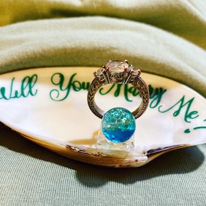 May include: A white seashell ring dish with the words "Will You Marry Me?" written in green calligraphy. A silver engagement ring with a large center stone and two smaller stones on either side is sitting on a blue glass orb inside the dish.