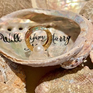 May include: A gold ring with the words "Will You Marry Me" inscribed on the inside of a seashell. The ring is sitting on a small white pearl.