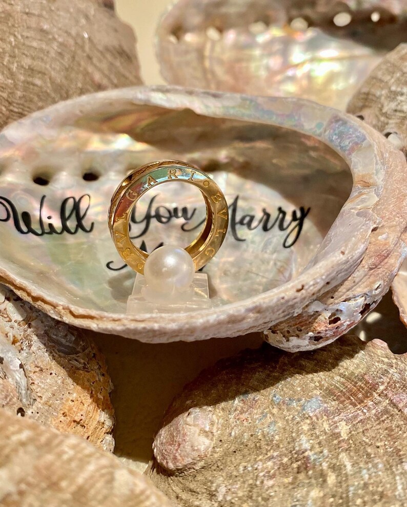 May include: A gold ring with the words "Will You Marry Me" inscribed on the inside of a seashell. The ring is sitting on a small white pearl.