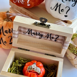 Puede incluir: Una caja de madera con el texto "Marry Me?" en su interior, que contiene una pequeña calabaza naranja con un anillo de diamantes. Dos calabazas más con "Marry Me?" están en el fondo. Las calabazas son naranjas y blancas.
