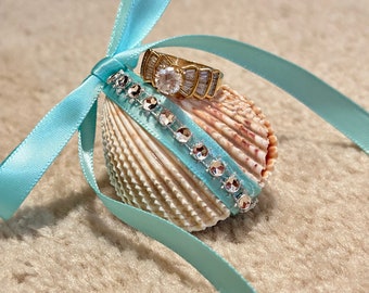 May include: A seashell ring holder with a light blue ribbon tied around it. The ring holder is decorated with rhinestones and a gold ring with a large clear stone is sitting on top.