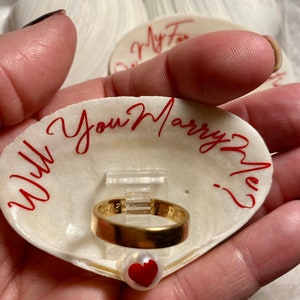 May include: A white seashell ring box with the words "Will you marry me?" written in red. A gold wedding band is displayed inside the box, resting on a small red heart.