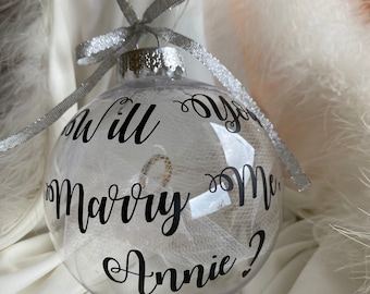 Op de afbeelding: Een transparante glazen kerstbal met een zilveren lint en de tekst "Will you marry me Annie?" in zwarte cursieve letters.