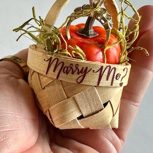 Puede incluir: Una pequeña cesta tejida con asa, que contiene una calabaza naranja en miniatura. Un anillo de oro con un diamante está colocado encima de la calabaza. La cesta tiene las palabras "Marry Me?" escritas en borgoña.