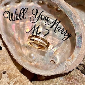 May include: A seashell ring holder with the words "Will You Marry Me?" written in black script. A gold ring is displayed in the holder.