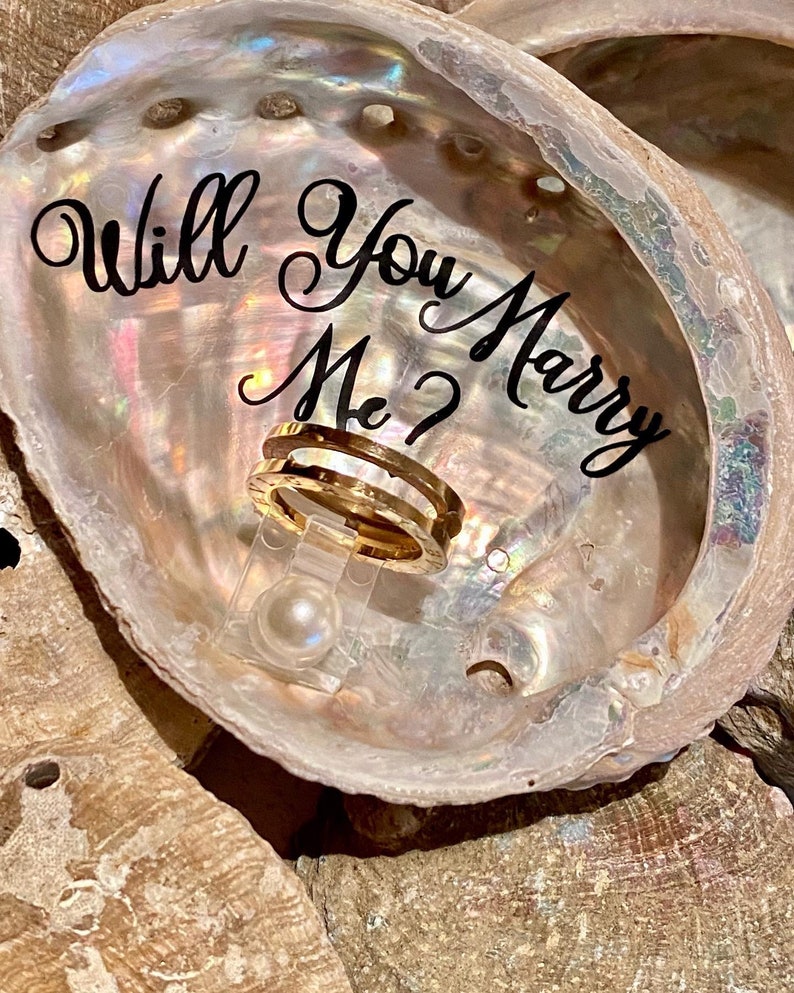 May include: A seashell ring holder with the words "Will You Marry Me?" written in black script. A gold ring is displayed in the holder.