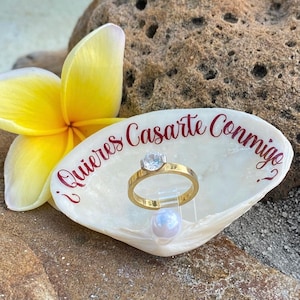 May include: A white seashell ring holder with the text "¿Quieres Casarte Conmigo?" in red lettering. A gold ring with a clear gemstone is displayed on the holder, along with a white pearl.
