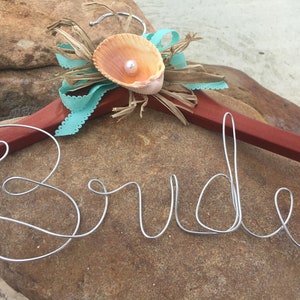 May include: A wooden hanger decorated with a seashell and pearl, with the word "Bride" written in wire.