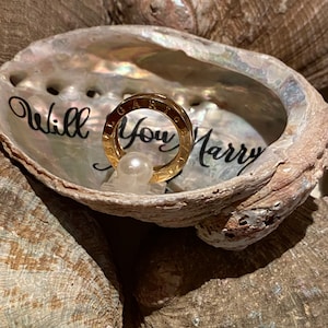 May include: A gold ring with the words "Will You Marry" engraved on it, sitting in a seashell with the same words written inside.